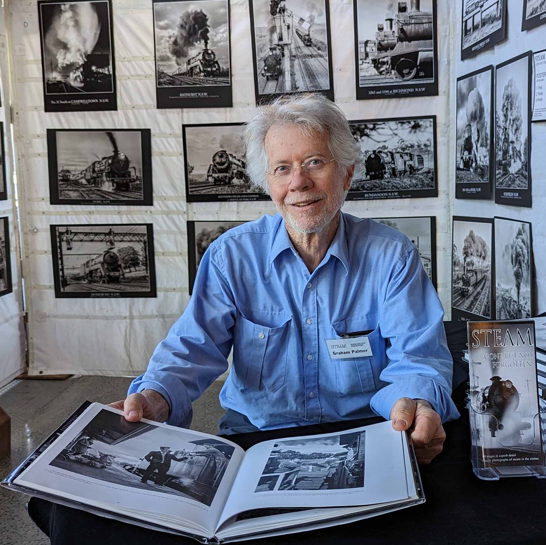 Graham Palmer seated at a table with his open book, framed steam-train prints displayed behind him.