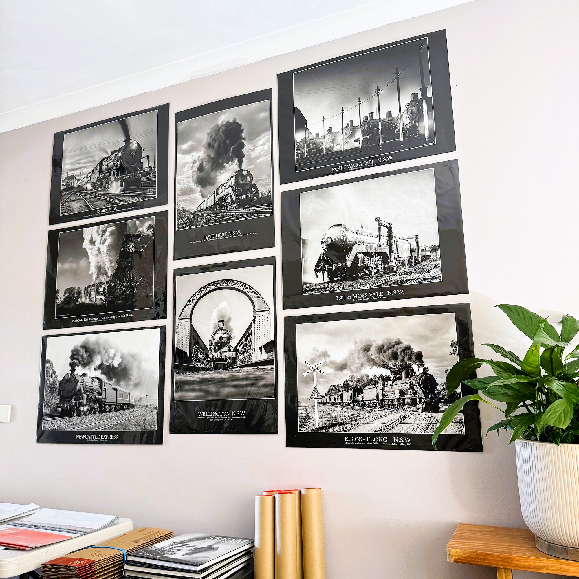 A wall of black-and-white steam-train prints from various NSW locations, with poster tubes on a table below.